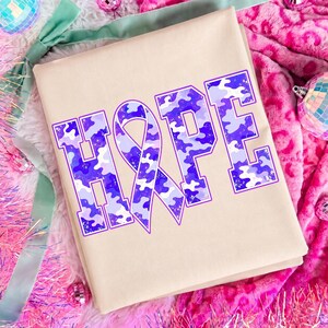 May include: A beige t-shirt featuring the word HOPE in large block letters. The letter O is replaced by an awareness ribbon. The text has a purple and light blue camouflage pattern with a distressed texture and a thin purple outline.