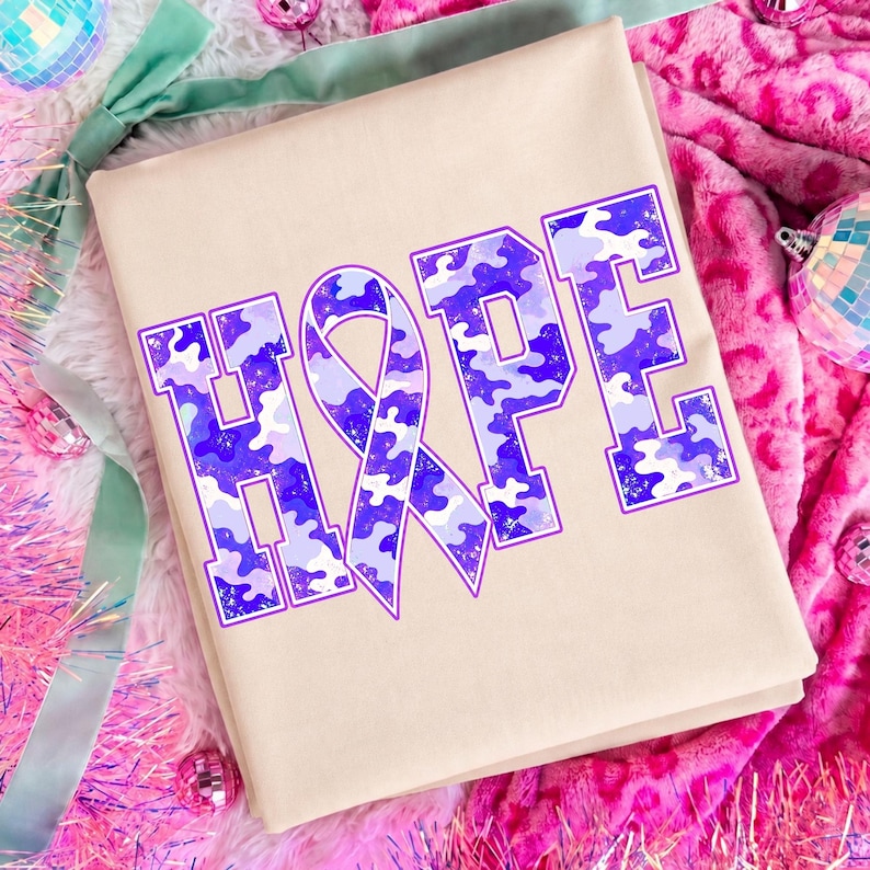 May include: A beige t-shirt featuring the word HOPE in large block letters. The letter O is replaced by an awareness ribbon. The text has a purple and light blue camouflage pattern with a distressed texture and a thin purple outline.