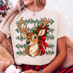 May include: A cream-coloured short-sleeve t-shirt featuring a graphic of a brown deer with large antlers. The deer is adorned with a bright red ribbon and green pine branches with red berries. The background includes horizontal pine branch patterns.