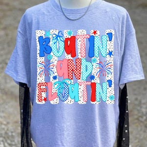 Op de afbeelding: Een lichtgrijs t-shirt met een grafische print met de tekst "BOATIN' AND FLOATIN'". Het ontwerp bevat verticale witte strepen met zwarte stippen, rood-blauwe ruitpatronen, vuurwerk en lichtblauwe strikjes op de letters.