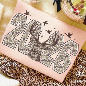 Puede incluir: Camiseta rosa claro con el año "2026" en un patrón de camuflaje y un gráfico de ciervo. El diseño incluye pájaros volando y estrellas. La camiseta se muestra sobre una tela con estampado de leopardo.