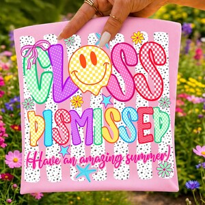 May include: A light pink fabric item featuring the text "CLASS DISMISSED" in colorful, checkered, and patterned bubble letters. A yellow checkered smiley face replaces the letter A. The design includes small flowers, stars, and "Have an amazing summer" text.