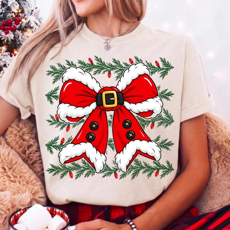 Puede incluir: Una camiseta de color crema con un gr&aacute;fico grande de un lazo rojo estilo Santa Claus con borde de piel blanca, un cintur&oacute;n negro con hebilla dorada y botones negros. El dise&ntilde;o est&aacute; rodeado de ramas de pino verdes y peque&ntilde;as luces rojas.