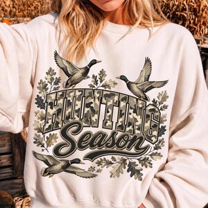 May include: A cream-colored crewneck sweatshirt featuring a graphic print with the text "HUNTING Season" in a camouflage pattern. The design includes three flying mallard ducks and oak leaves surrounding the bold, block-style lettering.