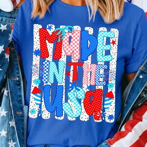 May include: A royal blue t-shirt featuring a graphic print with the text MADE IN THE USA. The design includes vertical white stripes with black dots, red and blue checkered patterns, light blue bows, small stars, and firework illustrations.
