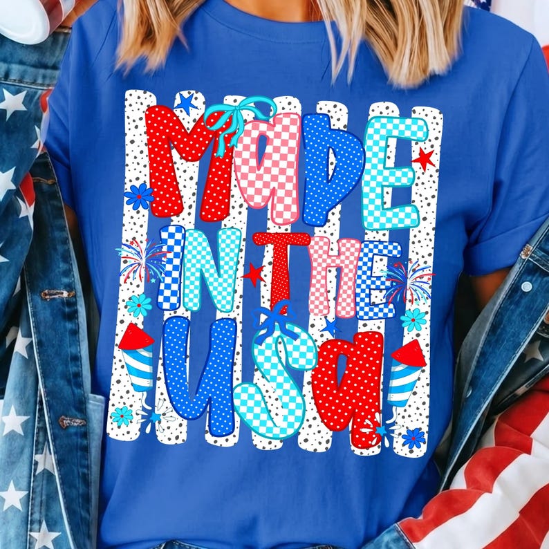May include: A royal blue t-shirt featuring a graphic print with the text MADE IN THE USA. The design includes vertical white stripes with black dots, red and blue checkered patterns, light blue bows, small stars, and firework illustrations.