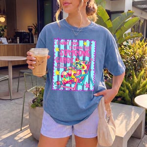K&ouml;nnte beinhalten: Ein blaues Kurzarm-T-Shirt mit einer Grafik eines bunten Waschb&auml;ren, der mit einem Getr&auml;nk in einem Liegestuhl sitzt. Der Text lautet I WANT YOU TO KNOW THAT SOMEONE CARES NOT ME BUT SOMEONE. Das Design enth&auml;lt pinke, gelbe und t&uuml;rkisfarbene Akzente.