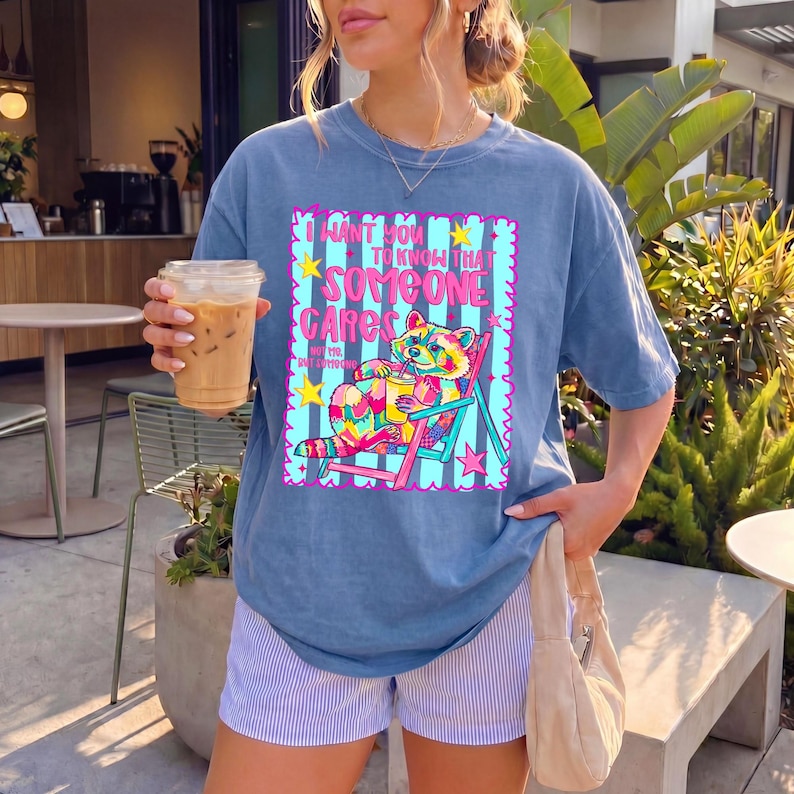 K&ouml;nnte beinhalten: Ein blaues Kurzarm-T-Shirt mit einer Grafik eines bunten Waschb&auml;ren, der mit einem Getr&auml;nk in einem Liegestuhl sitzt. Der Text lautet I WANT YOU TO KNOW THAT SOMEONE CARES NOT ME BUT SOMEONE. Das Design enth&auml;lt pinke, gelbe und t&uuml;rkisfarbene Akzente.