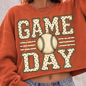 May include: A burnt orange cropped sweatshirt featuring a graphic print with the text GAME DAY in large, cream-coloured block letters with black polka dots. A detailed illustration of a baseball is positioned between the words GAME and DAY.