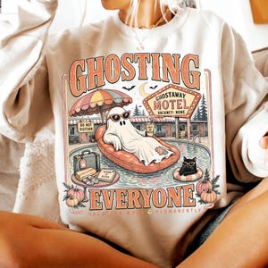 Puede incluir: Una sudadera beige de cuello redondo con un estampado gr&aacute;fico de un fantasma con gafas de sol en un flotador naranja, un gato negro, un letrero de motel que dice GHOSTAWAY MOTEL VACANCY: NONE y el texto GHOSTING EVERYONE VACATION MODE PERMANENTLY.