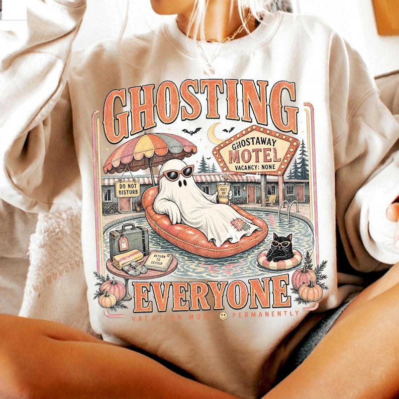 Puede incluir: Una sudadera beige de cuello redondo con un estampado gr&aacute;fico de un fantasma con gafas de sol en un flotador naranja, un gato negro, un letrero de motel que dice GHOSTAWAY MOTEL VACANCY: NONE y el texto GHOSTING EVERYONE VACATION MODE PERMANENTLY.