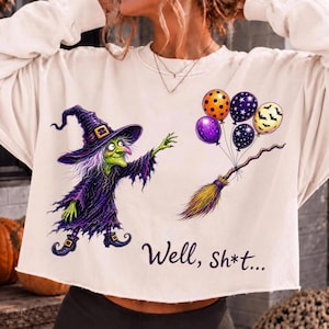 K&ouml;nnte beinhalten: Ein cremefarbenes Langarm-Sweatshirt mit einer Grafik einer gr&uuml;nh&auml;utigen Hexe in violettem Gewand und Hut, die nach einem Besenstiel mit vier bunten Luftballons greift. Der Text "Well, Sh*t..." ist darunter in Schwarz aufgedruckt.