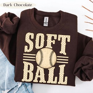 以下が含まれることがあります： ダークチョコレートブラウンのクルーネックトレーナー。ライトベージュのドット柄フォントで「SOFT BALL」とプリントされています。中央には野球ボールのグラフィックが配置され、ボールの両側には水平線が伸びています。