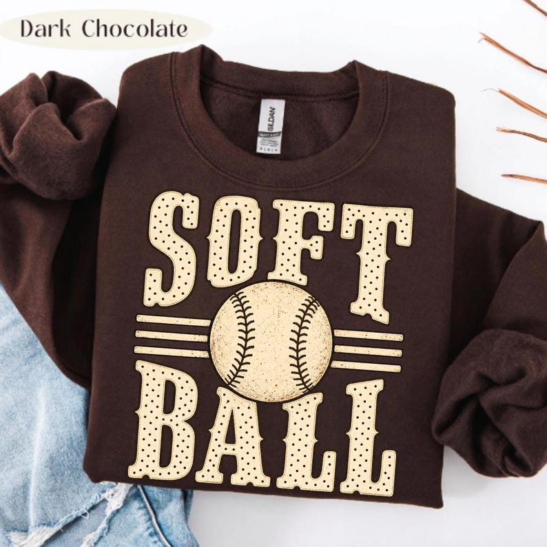 以下が含まれることがあります： ダークチョコレートブラウンのクルーネックトレーナー。ライトベージュのドット柄フォントで「SOFT BALL」とプリントされています。中央には野球ボールのグラフィックが配置され、ボールの両側には水平線が伸びています。
