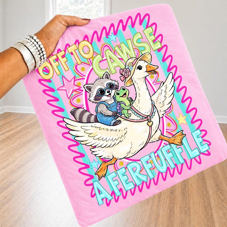 May include: Pink blanket with a cartoon illustration of a raccoon and a frog riding a goose. The blanket has the text "OFF TO CAWSE A FERFUFFLE" in a colorful, whimsical font. The blanket is held up by a person.