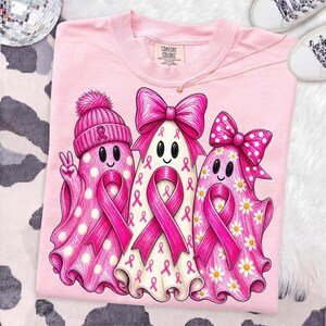Puede incluir: Una camiseta rosa claro con un gr&aacute;fico de tres fantasmas. El fantasma de la izquierda lleva un gorro rosa con un lazo, el central tiene un lazo rosa grande y el de la derecha un lazo rosa de lunares. Cada fantasma muestra un lazo rosa.