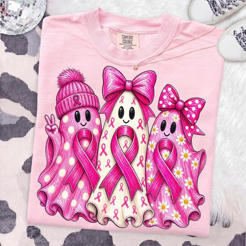 Puede incluir: Una camiseta rosa claro con un gr&aacute;fico de tres fantasmas. El fantasma de la izquierda lleva un gorro rosa con un lazo, el central tiene un lazo rosa grande y el de la derecha un lazo rosa de lunares. Cada fantasma muestra un lazo rosa.