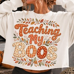 Pu&ograve; includere: Una felpa bianca a maniche lunghe con la scritta "Teaching My BOOS" in un carattere decorativo. Le lettere sono riempite con motivi floreali e a quadri arancioni. Il design &egrave; circondato da una corona di foglie e rami autunnali.