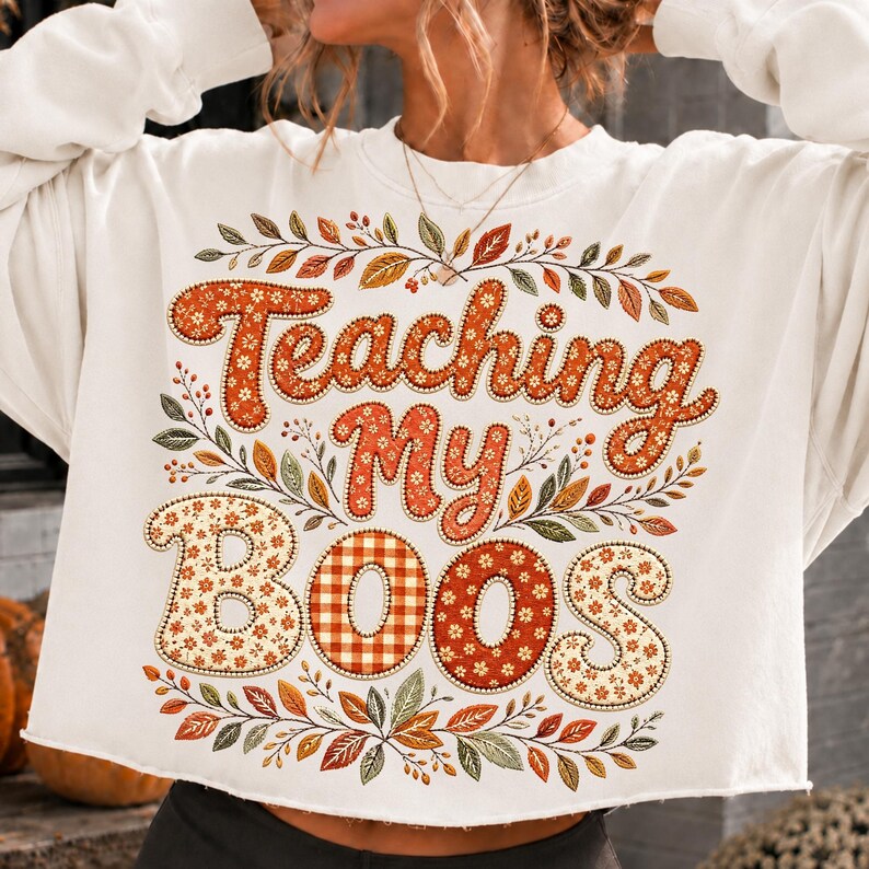 Pu&ograve; includere: Una felpa bianca a maniche lunghe con la scritta "Teaching My BOOS" in un carattere decorativo. Le lettere sono riempite con motivi floreali e a quadri arancioni. Il design &egrave; circondato da una corona di foglie e rami autunnali.