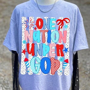 Pu&ograve; includere: Una t-shirt grigio chiaro con una stampa grafica che riporta il testo ONE NATION UNDER GOD in lettere con motivi rossi, bianchi e blu. Il design include strisce verticali punteggiate, fuochi d'artificio, stelle, un nastro rosso e razzi.
