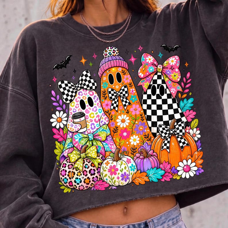 Puede incluir: Una sudadera corta de color gris carb&oacute;n con un gr&aacute;fico colorido de tres fantasmas decorados con patrones florales, estampado de leopardo y lazos a cuadros. El dise&ntilde;o incluye calabazas, margaritas y peque&ntilde;os murci&eacute;lagos decorativos.