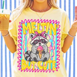 Puede incluir: Una camiseta de color amarillo p&aacute;lido con el gr&aacute;fico de un mapache gris que lleva una diadema rosa y gafas de sol en forma de coraz&oacute;n mientras sostiene un helado rosa derriti&eacute;ndose. El texto dice MELTIN' BUT CUTE en letras amarillas con borde rosa.