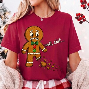 May include: A maroon t-shirt featuring a graphic of a brown gingerbread person with a worried expression, a green bow tie, and red and green buttons. The gingerbread person has a missing foot with crumbs, and the text reads well, shit...