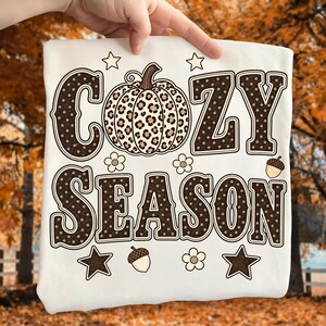 Może przedstawiać: Biały t-shirt z nadrukiem graficznym z napisem COZY SEASON wykonanym dużymi brązowymi literami w kropki. Wz&oacute;r zawiera centralnie umieszczoną dynię w panterkę, otoczoną małymi brązowymi gwiazdkami, kwiatami i ilustracjami żołędzi.