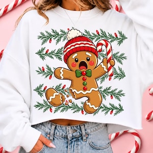 May include: A white cropped sweatshirt featuring a graphic of a surprised gingerbread person wearing a red and white knit hat and a green bow tie, holding a candy cane. The background print includes horizontal green pine branches and red lights.