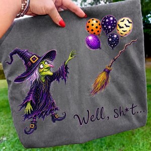 K&ouml;nnte beinhalten: Ein anthrazitfarbenes T-Shirt mit einer gr&uuml;nh&auml;utigen Hexe in violettem Gewand und Hut, die nach einem Besen greift, der an orangefarbenen, violetten und gelben Luftballons mit Stern- und Fledermausmustern befestigt ist. Der Text "Well, sh*t..." ist aufgedruckt.