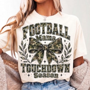 May include: A cream-coloured short-sleeve t-shirt featuring a graphic print with the text FOOTBALL MAMA TOUCHDOWN SEASON in a green camouflage pattern. The design includes a large central camouflage bow and decorative leaf accents on both sides.