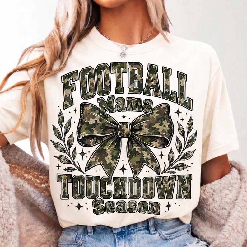May include: A cream-coloured short-sleeve t-shirt featuring a graphic print with the text FOOTBALL MAMA TOUCHDOWN SEASON in a green camouflage pattern. The design includes a large central camouflage bow and decorative leaf accents on both sides.