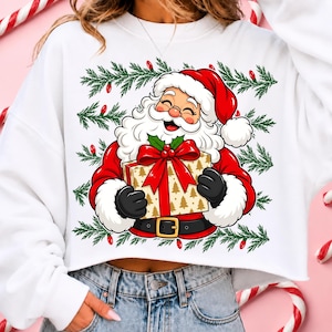 May include: A white long-sleeve sweatshirt featuring a graphic of Santa Claus wearing a red suit and hat, holding a gift box wrapped in gold paper with a large red bow. The design is surrounded by green pine branches and small red light bulbs.