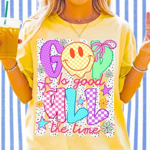 May include: A yellow t-shirt featuring a graphic print with the text "GOD Is good ALL the time". The design includes a yellow checkered smiley face, pink and blue checkered letters, pink bows, colorful stars, and small flowers on a dotted background.