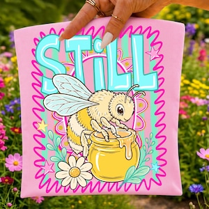 Puede incluir: Un art&iacute;culo de tela rosa claro con un gr&aacute;fico de una abeja peluda posada sobre un tarro de miel amarillo. La palabra STILL est&aacute; impresa en letras grandes de color azul claro sobre la abeja. El dise&ntilde;o incluye una margarita y estrellas.