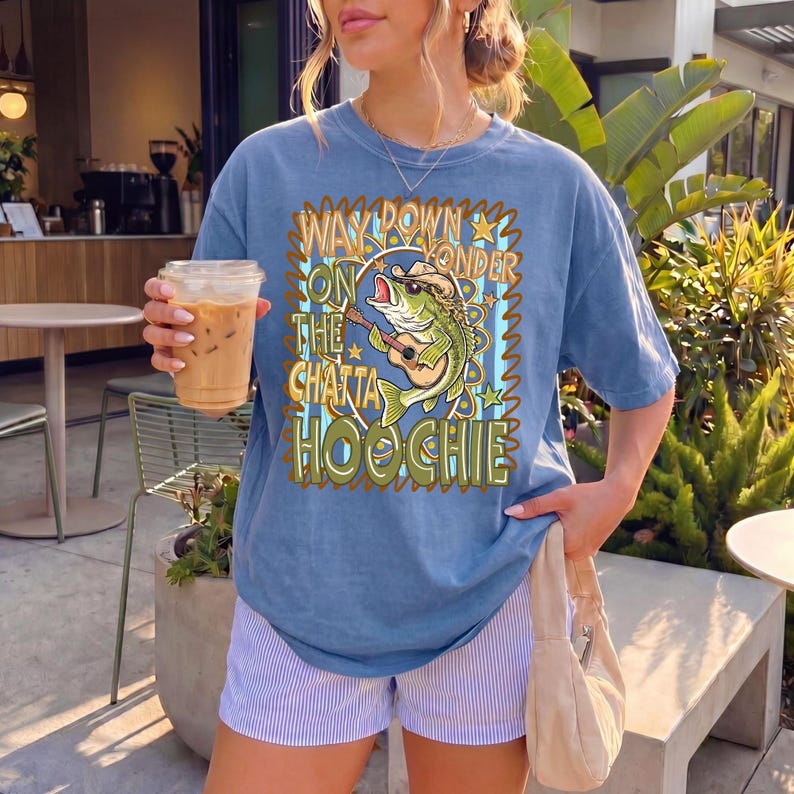 May include: A blue t-shirt with the text "Way Down on the Chatta Hoochie" and a graphic of a fish playing a guitar. The shirt is paired with striped shorts and a beige purse. The person is holding an iced coffee.