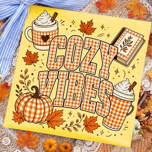 Puede incluir: Un letrero cuadrado amarillo con el texto COZY VIBES en estampado de cuadros vichy naranja y blanco. El dise&ntilde;o incluye ilustraciones de una calabaza, dos tazas de chocolate caliente con nata y un libro, rodeados de hojas de arce oto&ntilde;ales.
