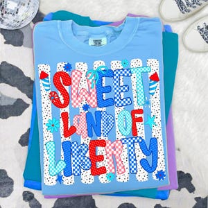 K&ouml;nnte beinhalten: Ein hellblaues T-Shirt mit dem Text SWEET LAND OF LIBERTY in einem rot-wei&szlig;-blauen Karomuster. Das Design enth&auml;lt vertikale schwarze Punkte-Streifen, kleine blaue Blumen, Sterne und Feuerwerksgrafiken auf der Vorderseite.
