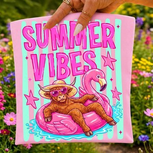 Pu&ograve; includere: Un articolo in tessuto rosa con il testo SUMMER VIBES in lettere rosa. Il design mostra una mucca highland marrone con cappello di paglia e occhiali da sole rosa, sdraiata su un salvagente a forma di fenicottero rosa, circondata da stelle rosa.