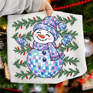 May include: A white t-shirt featuring a graphic of a snowman made of iridescent, multi-colored square tiles. The snowman wears a matching hat and scarf, with green pine branches and small red Christmas lights in the background.