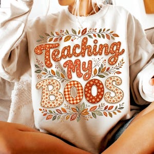 Pu&ograve; includere: Una felpa girocollo color crema con la scritta "Teaching My BOOS" in grandi lettere a fantasia. Il design &egrave; circondato da una ghirlanda decorativa di foglie dai colori autunnali nelle tonalit&agrave; dell'arancione, del marrone e del verde.
