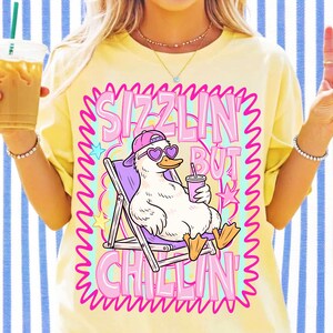 May include: A pale yellow t-shirt featuring a graphic of a white duck wearing a pink baseball cap and heart-shaped sunglasses. The duck sits in a purple lounge chair holding a drink. The text SIZZLIN' BUT CHILLIN' surrounds the duck in pink.