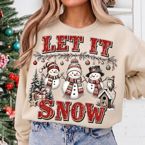 Pu&ograve; includere: Una felpa beige a girocollo con una stampa grafica che riporta il testo LET IT SNOW in rosso e verde. Il design include tre pupazzi di neve con sciarpe e cappelli, un piccolo gufo, una casetta per uccelli, un abete e un pacco regalo.