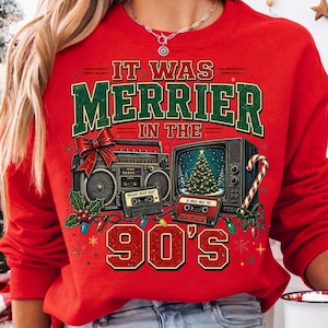 Może przedstawiać: Czerwona bluza z długim rękawem z nadrukiem graficznym i napisem IT WAS MERRIER IN THE 90'S. Wz&oacute;r przedstawia boombox, telewizor z choinką, dwie kasety magnetofonowe, czerwoną kokardę, ostrokrzew oraz laskę cukrową.