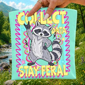 Op de afbeelding: Een licht mintgroene draagtas met een centrale illustratie van een grijze en witte wasbeer die een rock-and-roll handgebaar maakt. De tekst luidt COLLECT DATA bovenaan en STAY FERAL onderaan in dikke gele letters met roze accenten.