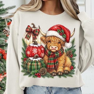 May include: A cream-colored crewneck sweatshirt featuring a graphic of a fluffy Highland cow wearing a red Santa hat and a green and red plaid scarf, surrounded by three pumpkins, pine cones, holly berries, and evergreen branches.