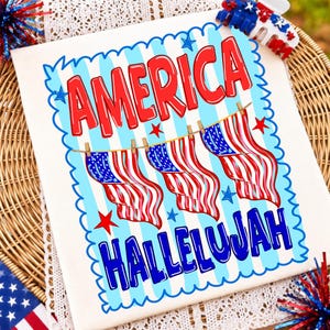 Puede incluir: Una camiseta blanca con un gr&aacute;fico que presenta el texto rojo AMERICA en la parte superior y el texto azul HALLELUJAH en la parte inferior. Entre las palabras hay tres banderas estadounidenses colgadas de un tendedero, con un borde a rayas azul.