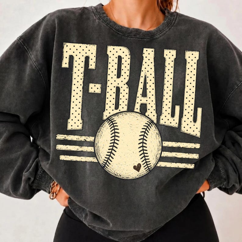 Pode incluir: Uma camisola de gola redonda em cinzento-carv&atilde;o com efeito lavado, apresentando o texto T-BALL numa fonte grande de cor creme com bolinhas. Abaixo do texto, encontra-se um gr&aacute;fico de uma bola de basebol com costuras e um pequeno cora&ccedil;&atilde;o.