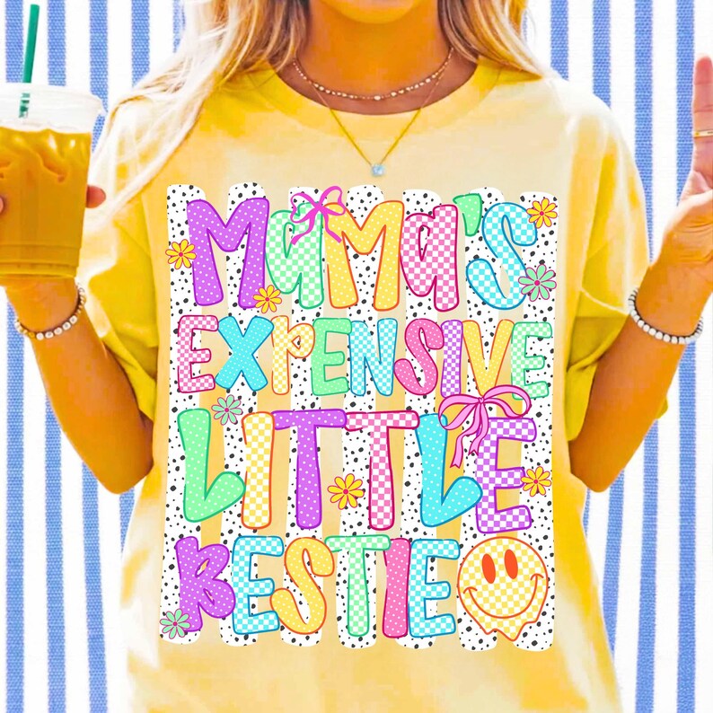 May include: A yellow short-sleeve t-shirt featuring a graphic print with the text "MAMA'S EXPENSIVE LITTLE BESTIE" in colorful, checkered, and polka-dot patterns. The design includes small flowers, pink bows, and a yellow smiley face icon.