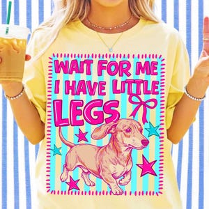 May include: A pale yellow t-shirt featuring a graphic print with a light blue and yellow striped background. The text reads "WAIT FOR ME I HAVE LITTLE LEGS" in bold pink letters, accompanied by a pink bow and pink stars around a brown dachshund.